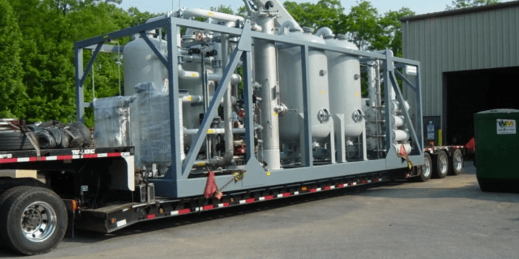 A large gray shipping container base with pipes and machinery on top sits on the back of a semi-truck.